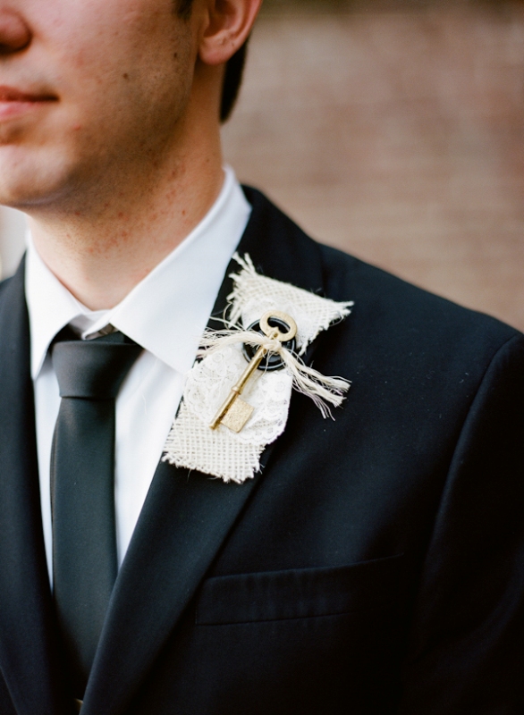 an unusual wedding boutonniere with white burlap, some printed fabric and a button and a large key shows off something personal for the groom