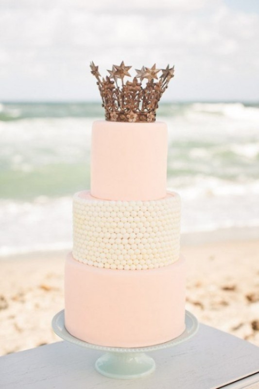 a light pink and white three tier wedding cake topped with a crown cake topper is a great fit for a royal wedding