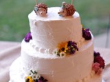 a white buttercream wedding cake decorated with bold fresh blooms and topper with hedgehog cake toppers for a woodland wedding