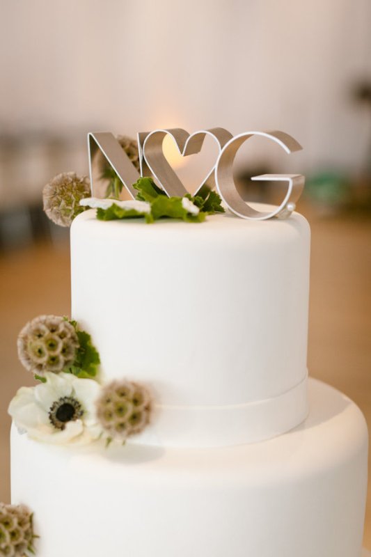 a pure white wedding cake with a ribbon, greenery, a white anemone, seed pods and metallic monogram cake toppers