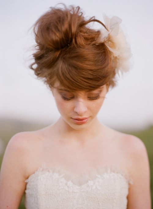 a messy wavy updo with bangs and a large faux flower for a modern bride with a touch of romance