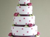 a white wedding cake with pink polka dots, black and pink ribbon, pink sugar blooms and leaves, a monogram cake topper