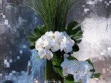 a tropical beach wedding centerpiece with tall glasses with floating candles, white orchids and monstera leaves plus grasses