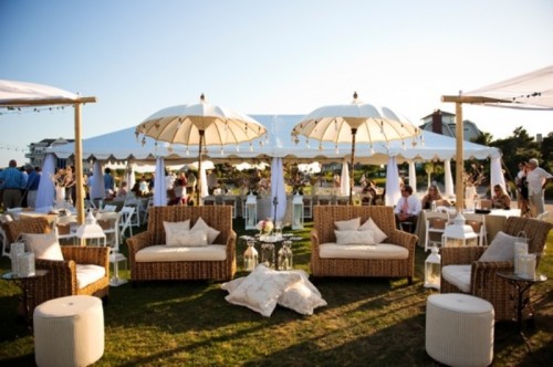 a white beach wedding lounge with wicker furniture and white upholstery, white poufs and umbrellas