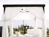 a chic white lounge with a white sofa, a coffee table and a cabana over them with white curtains plus a sea view is amazing