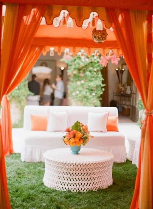 a bright outdoor wedding lounge with a white sofa and orange pillows, orange curtains and a chandelier, some bright blooms for a colorful summer wedding
