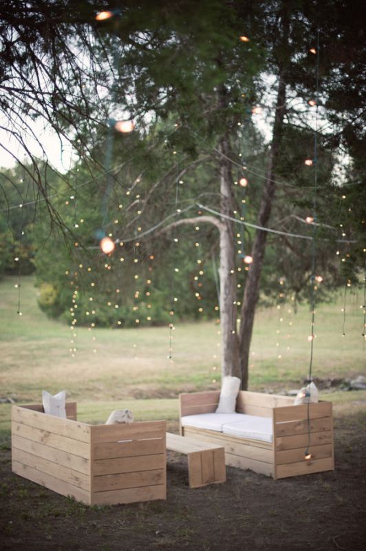 A simple outdoor wedding lounge with planked neutral furniture, white upholstery and lights over the space, hanging down from the tree