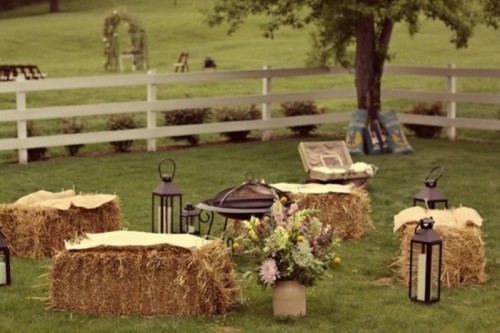 a rustic outdoor wedding lounge with hay stacks instead of sofas, a fire pit and candle lanterns and neutral blooms around