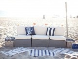 a small outdoor beach wedding lounge with a wicker sofa with neutral upholstery and navy pillows and rugs, towels, an umbrella over the sofa
