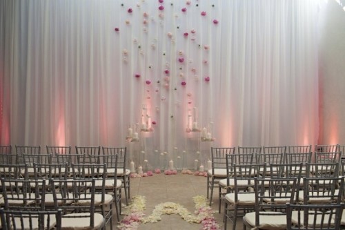 a white curtain with bright pink and neutral blooms attached, with candles on stands and on the floor plus more blooms