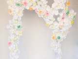 a wedding arch made of white, green, yellow and red paper flowers and featuring a creative shape