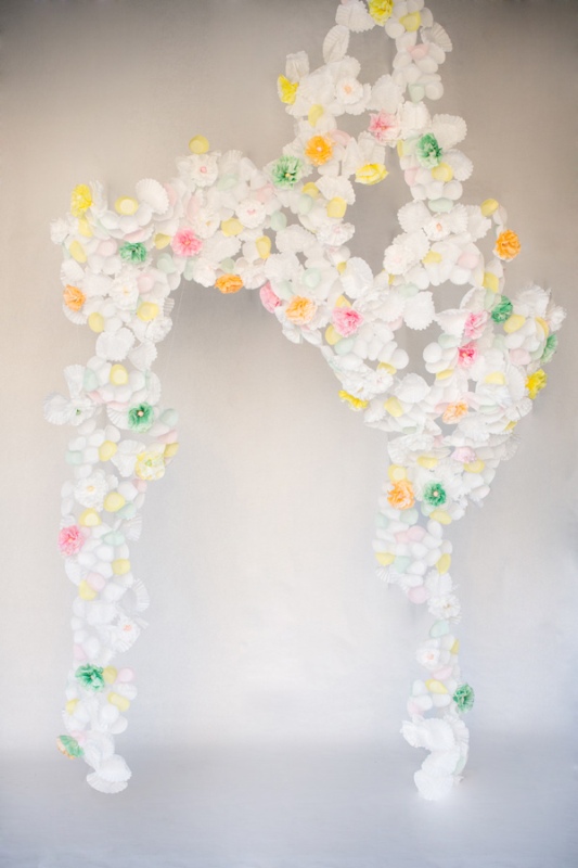 A wedding arch made of white, green, yellow and red paper flowers and featuring a creative shape