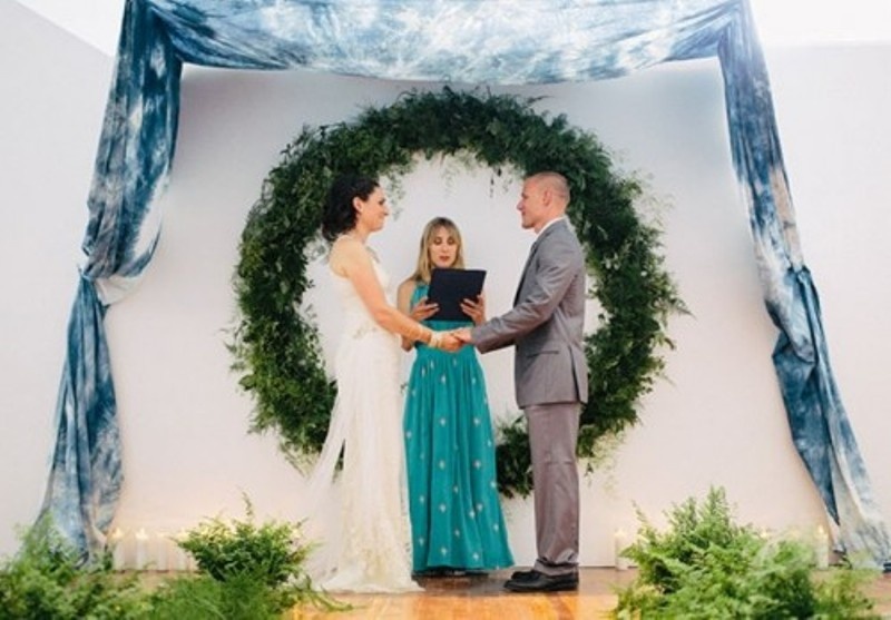 A round greenery wedding arch plus some tie dye blue fabric over will bring an outdoor feel indoors and will accent the couple