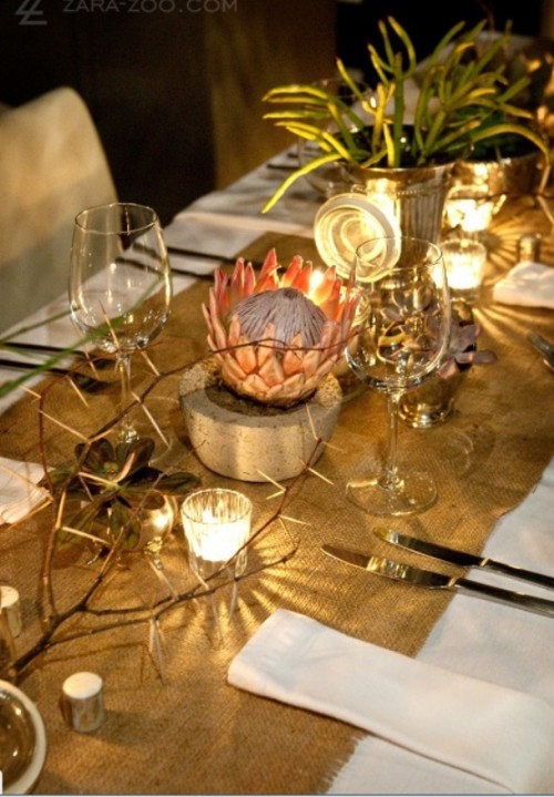 a safari-themed wedding table with a burlap runner, candles and a king protea in a concrete planter, greenery and twigs, simple cutlery