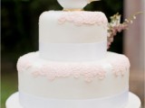 a white wedding cake with white ribbons, pink lace and some fresh blooms in a cup on top