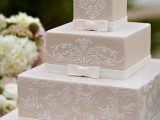 a square tan wedding cake decorated with white lace, ribbons and bows plus a sugar bloom on top