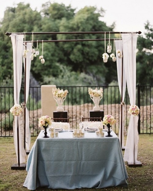 a sweetheart table decorated with lush white florals, candles and with a wedding arch with fabric, blooms and suspended candles