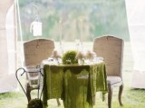 a sweetheart table topped with a green asymmetrical tablecloth, greenery, neutral blooms and an oversized candle holder next to the table