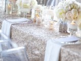 a silver glitter wedding tablecloth with white and pastel blooms, with candles and silver chargers is a very glam idea