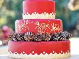 a red Christmas wedding cake with a patterned tier and paintings on it, with pinecones is a pretty and bold dessert