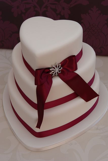 a refined white heart-shaped wedding cake with red ribbons and a bow with embellishments is a stylish and beautiful idea