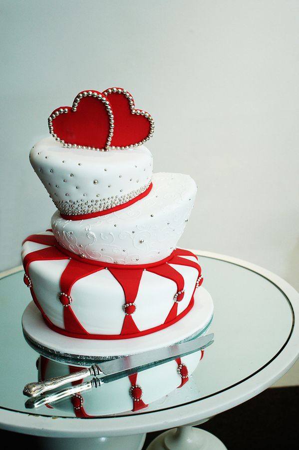 A whimsical and quirky white and red wedding cake with irregular tiers, embellishments and red heart and bead toppers is bold and gorgeous