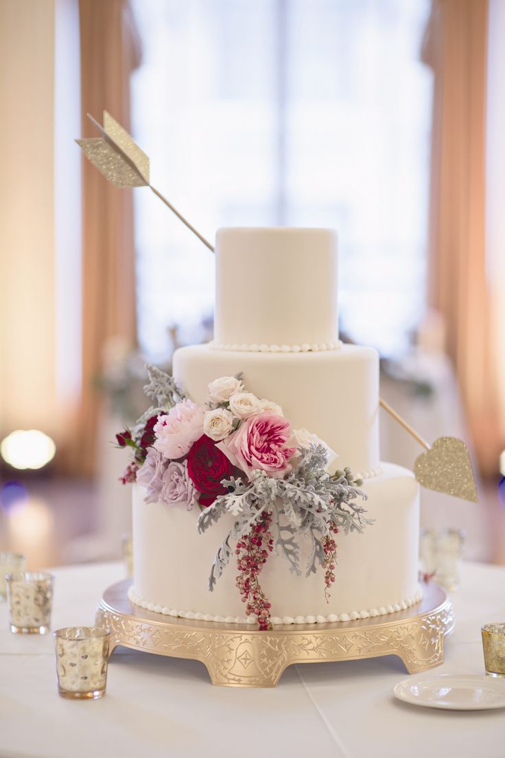 A beautiful Valentine wedding cake in white, with bold blooms and pale greenery plus an arrow with gold glitter feels very Valentine like