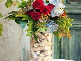 a tall clear vase filled with corks and with an arrangement of greenery, pale succulents and red flowers is extra bold