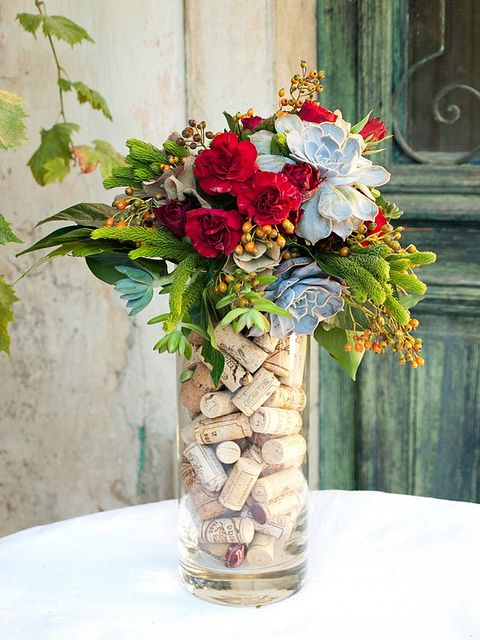 a tall clear vase filled with corks and with an arrangement of greenery, pale succulents and red flowers is extra bold