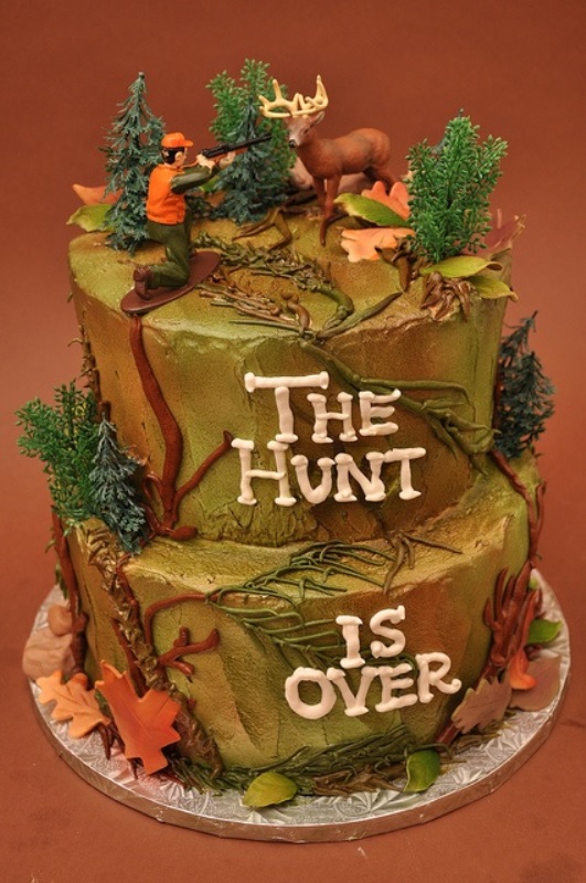 A bright wedding cake with sugar leaves, trees, a deer and a hunter on top hinting on a funny joke