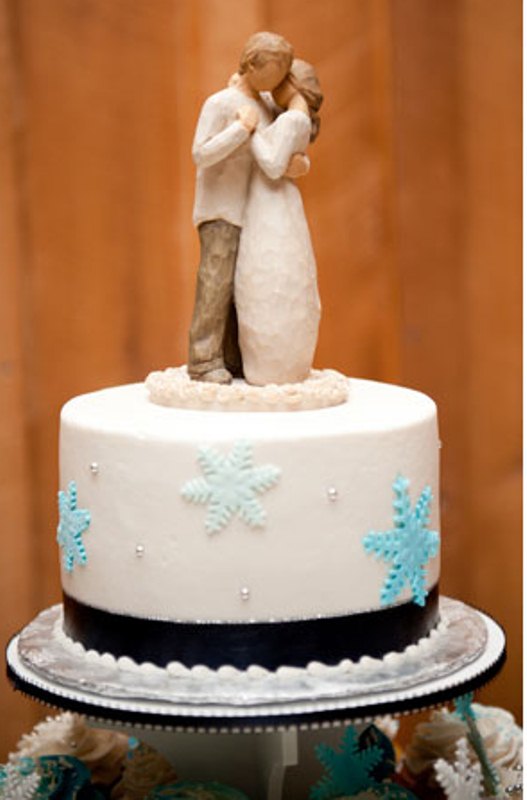A white wedding cake with black ribbon and blue sugar snowflakes plus a couple topper