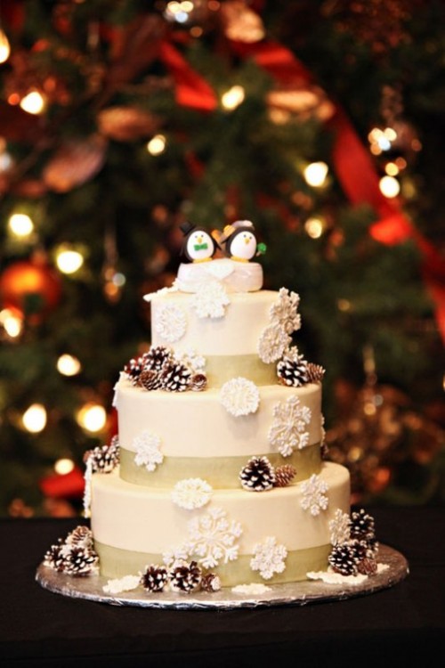 a white wedding cake with burlap ribbon, with snowy pinecones and snowflakes plus penguin cake toppers