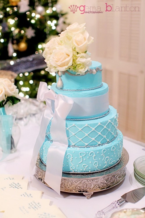 a turquoise wedding cake with patterns, a white ribbon bow and white roses on top