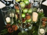 a shiny and cozy Christmas wedding centerpiece of a glass vase filled with green ornaments and pinecones, candles and fir branches on the table