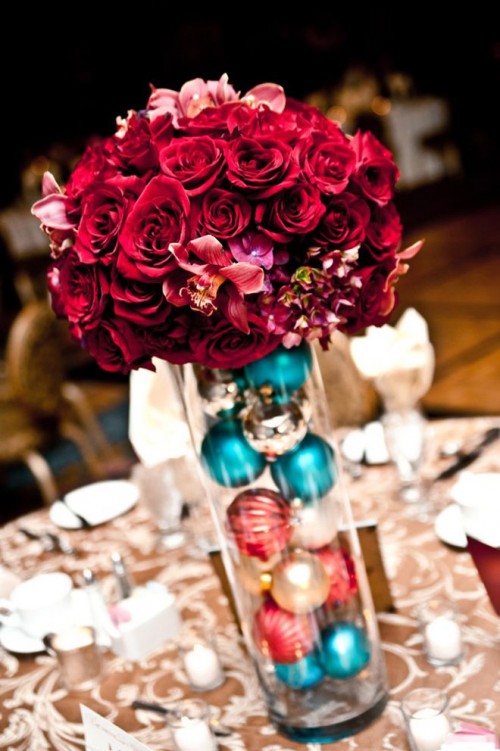 a colorful Christmas wedding centerpiece of a tall vase with colorful ornaments and bold burgundy blooms is a fun and quirky idea to try