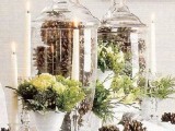 a refined Christmas wedding centerpiece of tall glass jars filled with pinecones, greenery and white blooms in bowls and pinecones on the table