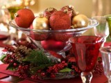 a Christmas wedding centerpiece of a glass bowl filled with burgundy and bold ornaments, with fir branches, berries and pinecones around