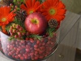 a rustic and chic Christmas wedding centerpiece of a glass bowl with berries, fir branches, red blooms, apples is a cozy idea for a farmhouse wedding