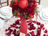 a Christmas wedding centerpiece of a tall red vase with bold blooms, greenery and glitter ornaments and red petals on the table will make a statement
