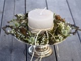 a chic rustic Christmas wedding centerpiece of a metallic stand with greenery, moss, thistles, pinecones and a candle in the center is a very cozy farmhouse piece