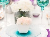 a bright wedding tablescape with turquoise glasses and glass plates, white, lilac and purple blooms, purple napkins and white roses is chic