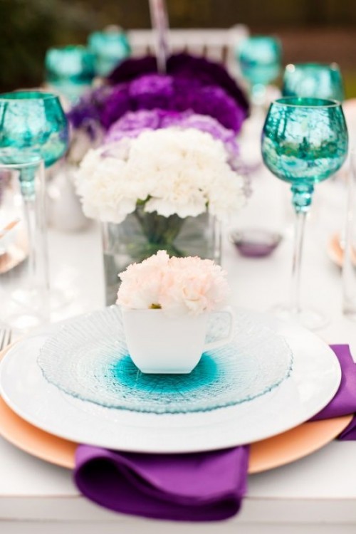 a bright wedding tablescape with turquoise glasses and glass plates, white, lilac and purple blooms, purple napkins and white roses is chic