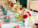 a bright wedding tablescape with pink napkins, blue containers with favors, pink, green and deep red blooms, turquoise napkins and placemats