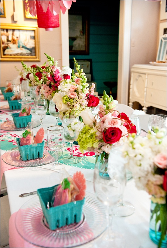 A bright wedding tablescape with pink napkins, blue containers with favors, pink, green and deep red blooms, turquoise napkins and placemats