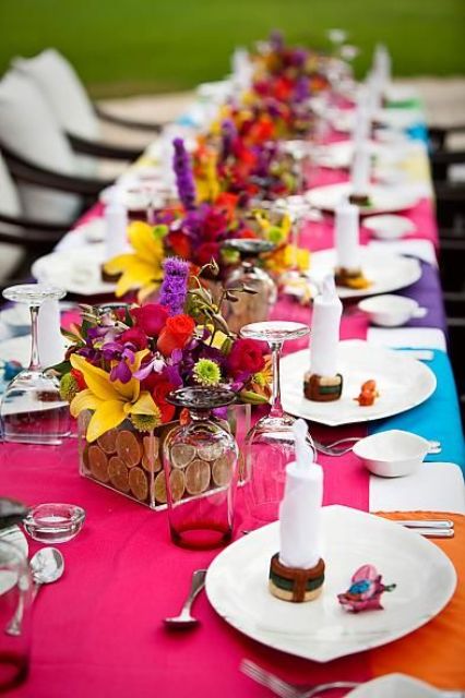 a jewel-tone wedding tablescape with a pink runner, colorful placemats, bold red, hot pink, yellow, green and purple blooms, candles and simple cutlery