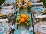 a bright wedding tablescape with a blue table runner and patterned plates, pink and yellow blooms and greenery, citrus in a box and candles is cool