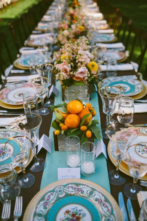 a bright wedding tablescape with a blue table runner and patterned plates, pink and yellow blooms and greenery, citrus in a box and candles is cool