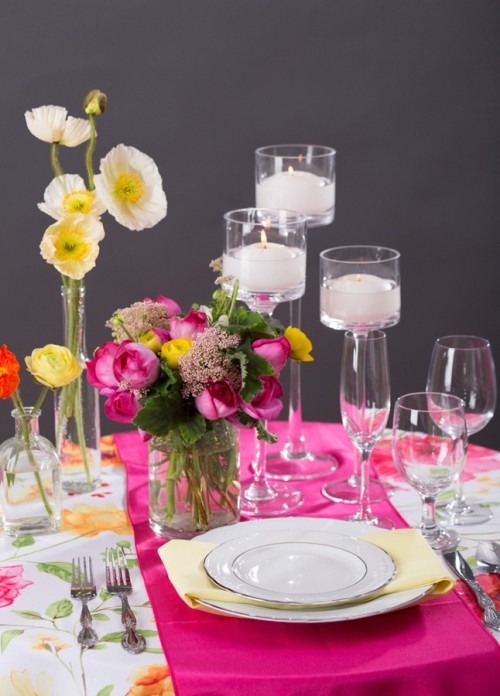 a hot pink place setting with a yellow napkin, hot pink and yellow blooms and candles in tall candleholders plus a floral tablecloth