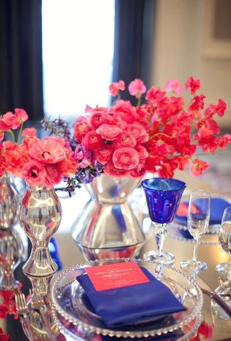 a bright wedding tablescape with hot pink and red blooms, an electric blue napkin and glasses, clear plates and mercury glass vases is amazing
