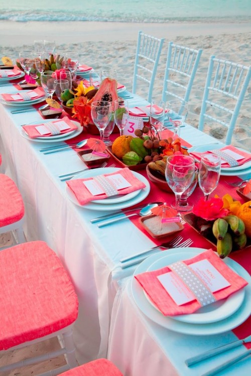 a bold tropical wedding tablescape with a blue tablecloth and plates, blue and coral chairs, a coral table runner and napkins, fruit centerpieces